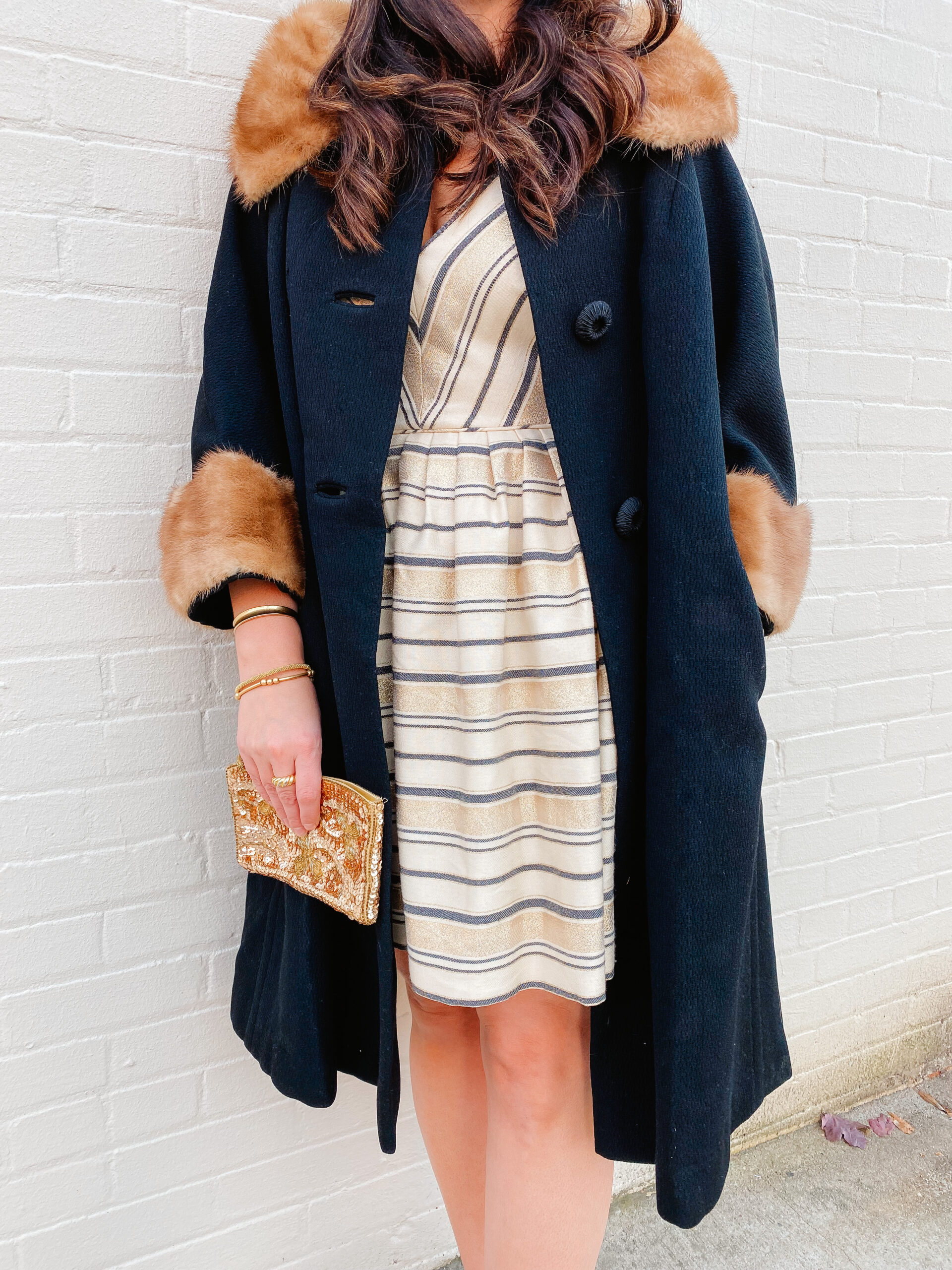 Emily Soto in a cream colored dress and faux fur lined jacket holding a gold clutch purse